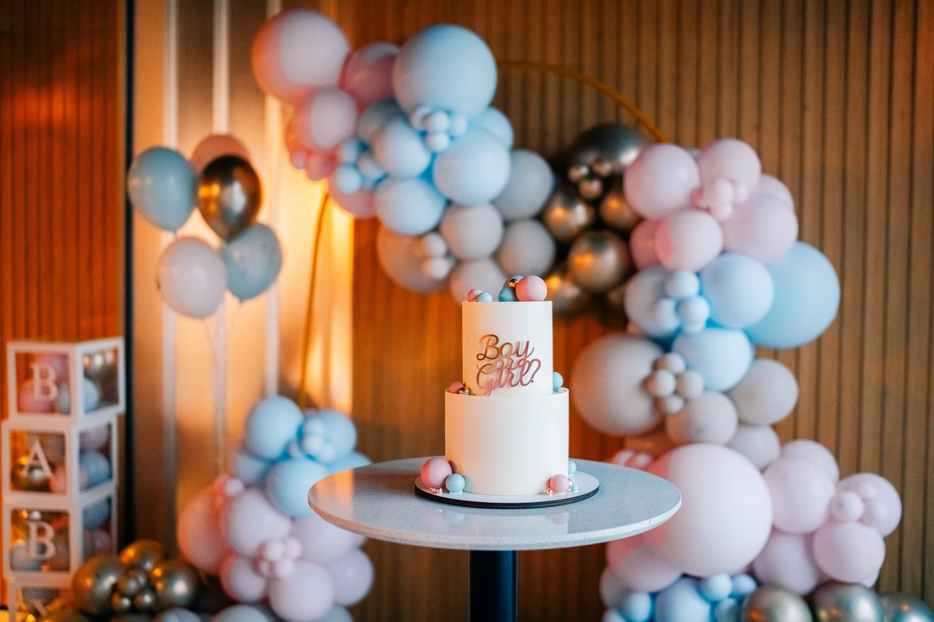 A baby shower is taking place with a stunning cake at the focal point, surrounded by pastel balloon decorations in pink and blue. The atmosphere is festive and cheerful.