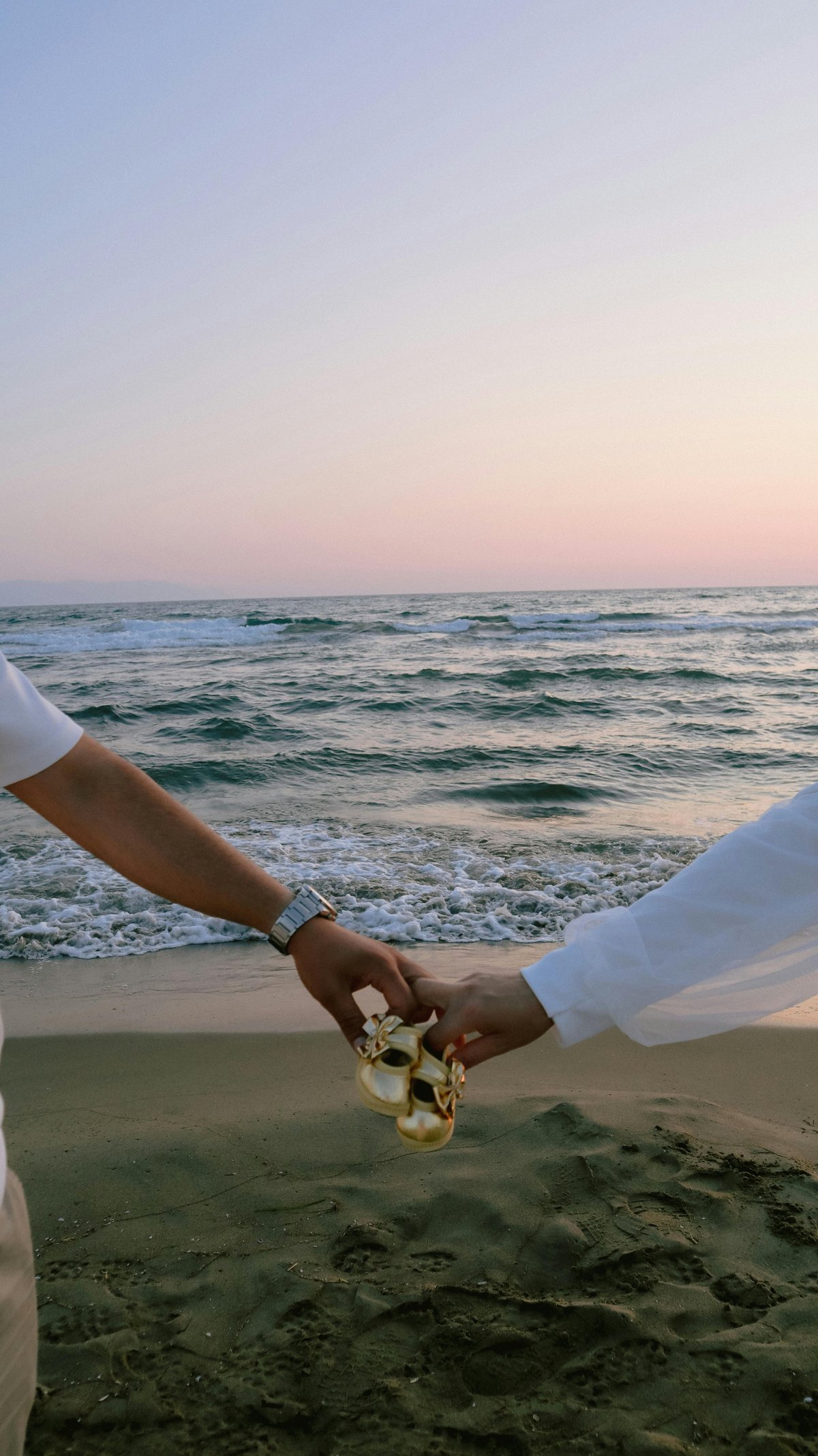 Couple holding hands on beach at sunset, wearing white clothing, with ocean waves and pink sky in background