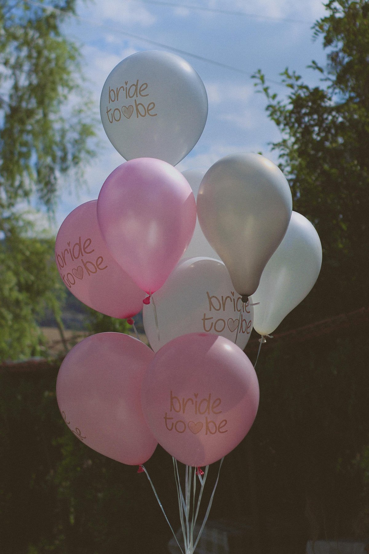 Cluster of pink and white balloons with bride to be text against blue sky and green foliage