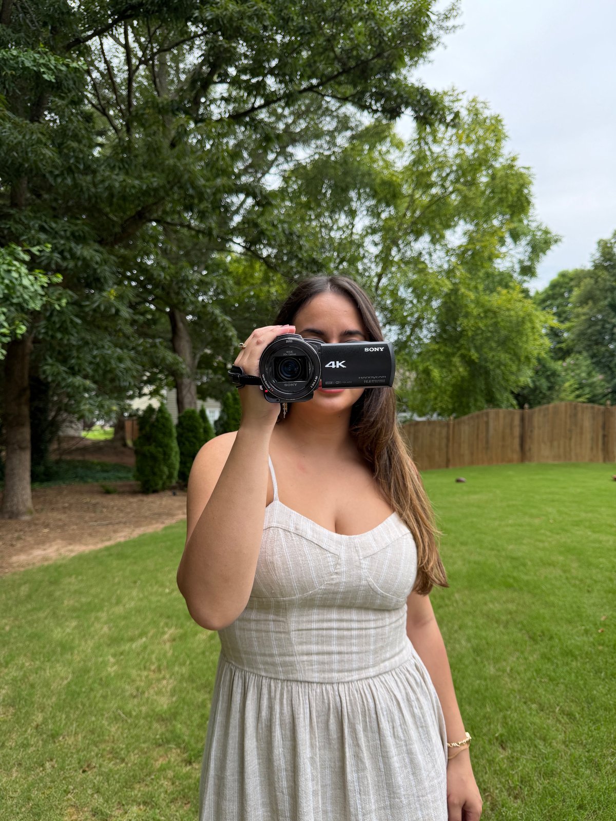 Woman in light dress holding 4K video camera to her face in a park with trees and green lawn