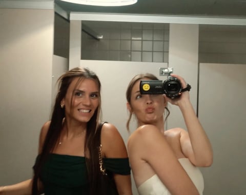 Two women taking a selfie in a bathroom mirror; one wearing black holds a camera while smiling at the camera