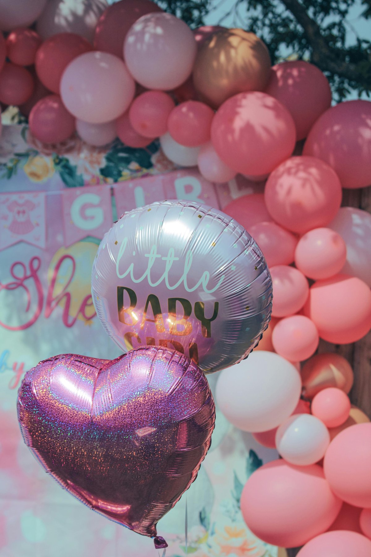 Baby shower decoration with metallic heart and round foil balloons in pink, white, and gold tones arranged above colorful banner