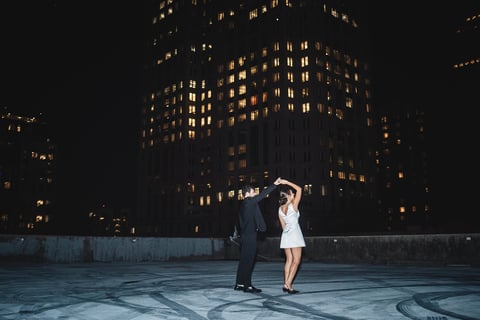 Man in suit and woman in white dress dancing on rooftop at night with illuminated city buildings in background
