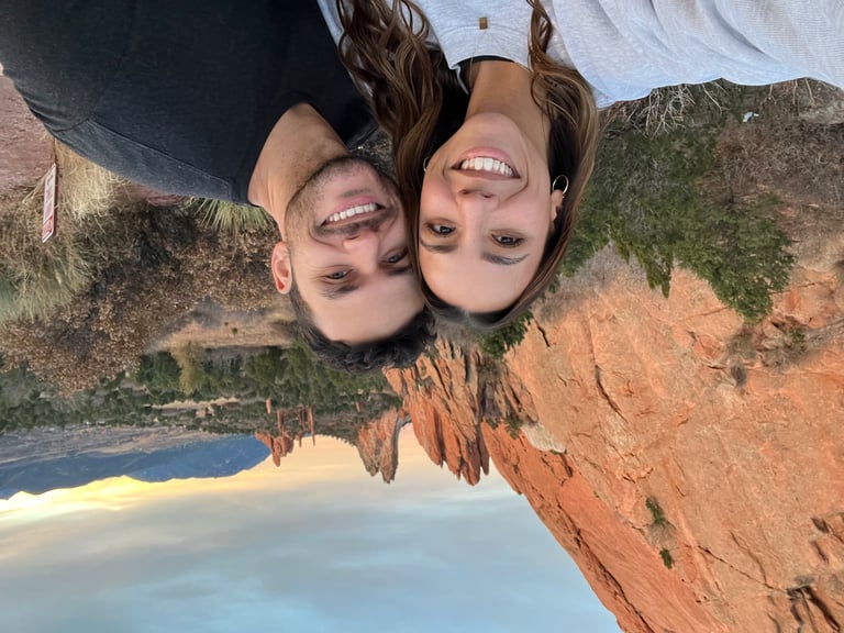 Two smiling people upside-down at a scenic overlook with red cliffs, ocean, and blue sky in background