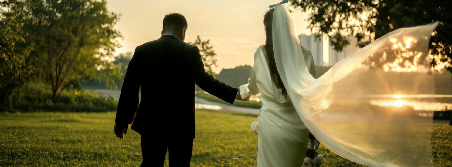 Bride and groom walking hand in hand across a sunny park with city skyline in background