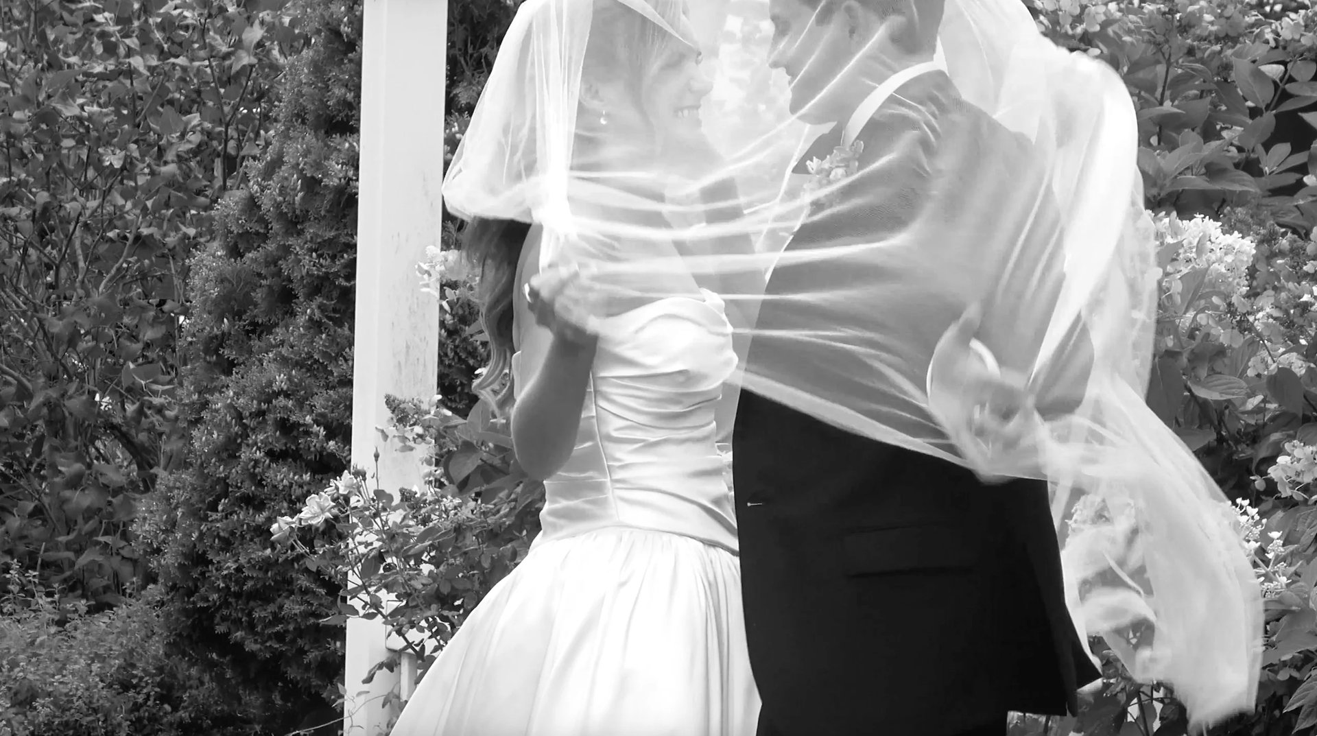 Black and white photo of bride and groom embracing, brides veil flowing in wind near white column