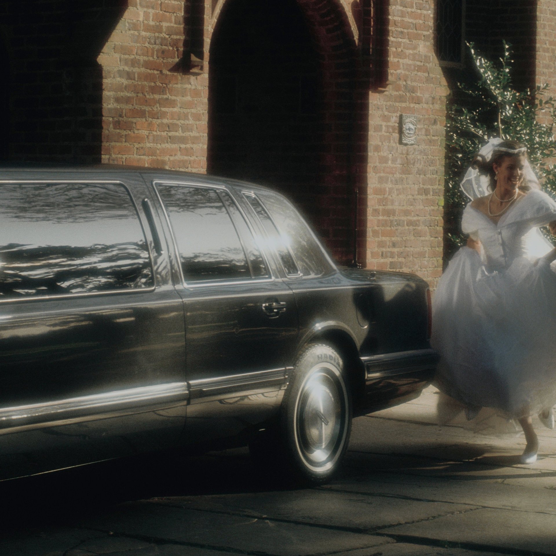 Woman in white wedding dress standing beside a black limousine in front of a brick building