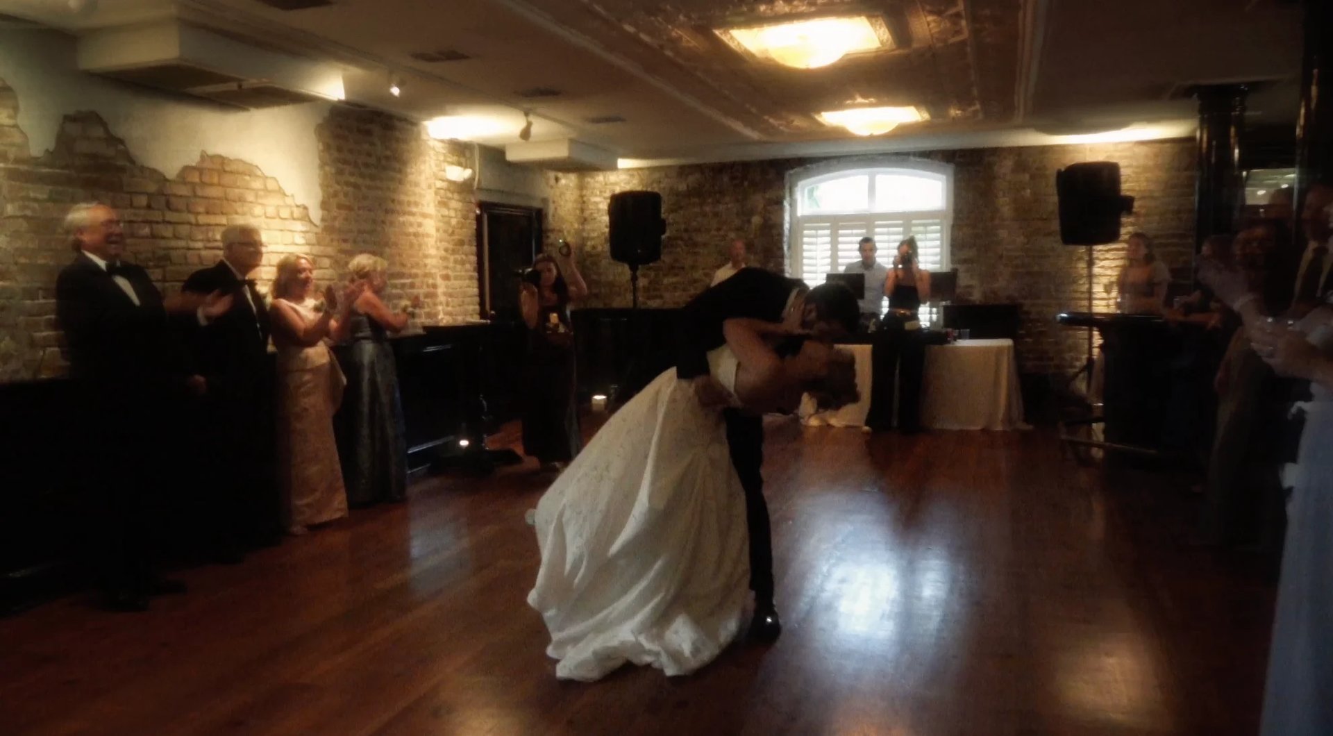 Bride and groom dancing at their wedding reception in a brick-walled venue with guests watching from the sides.