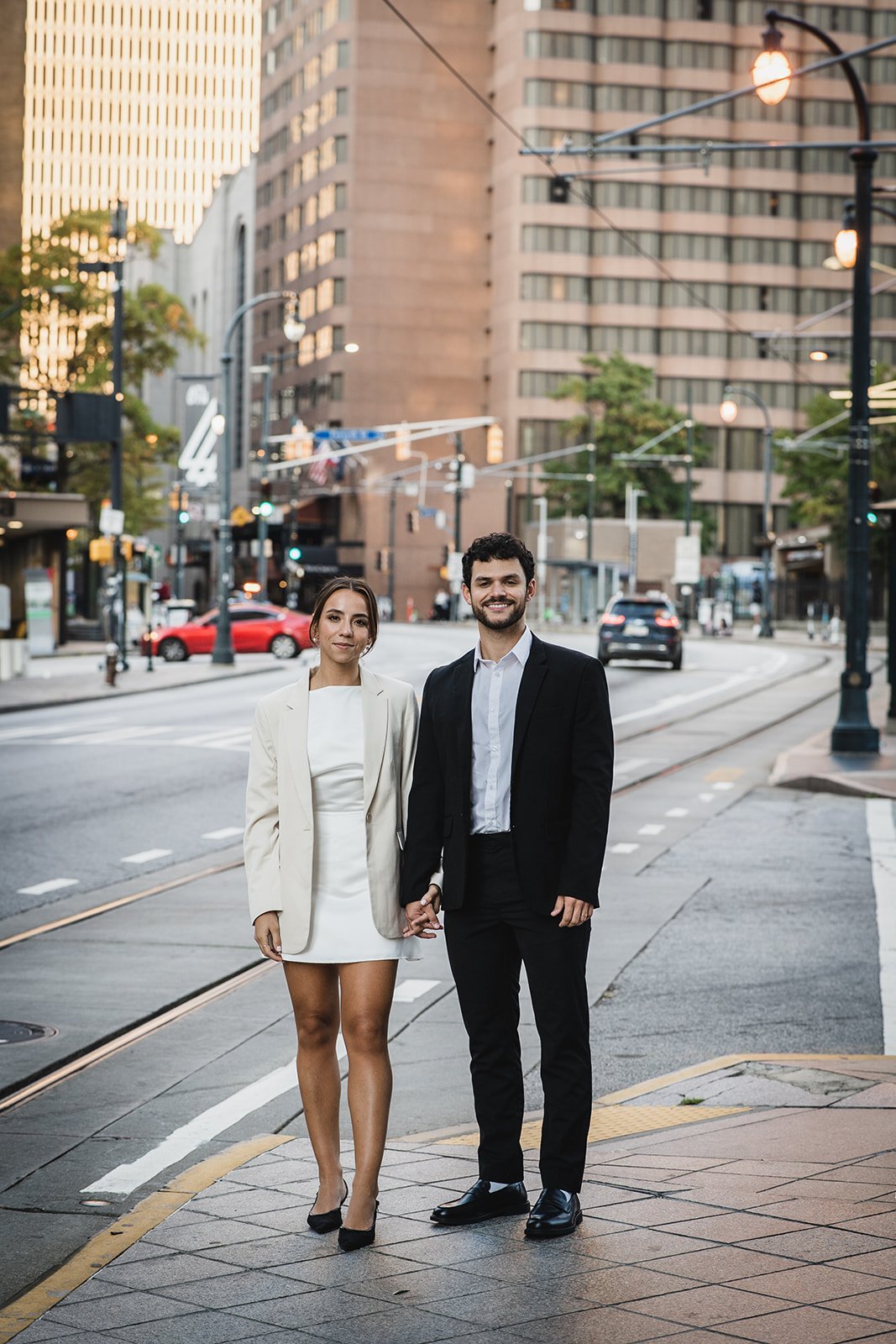 A well-dressed couple holding hands while standing on a city street surrounded by tall buildings and streetlights
