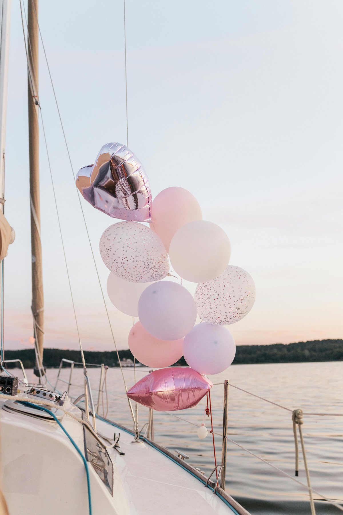 Cluster of pink and white balloons tied to the deck of a sailboat on calm water at sunset