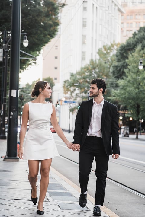 Couple in formal attire walking hand-in-hand down a city street with tall buildings and trees in the background