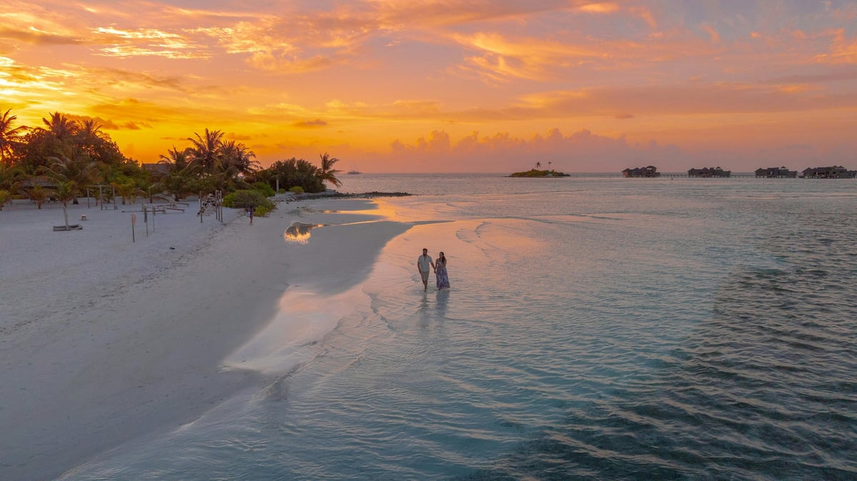 Couple wading in shallow turquoise water at sunset on a tropical beach with palm trees and distant boats