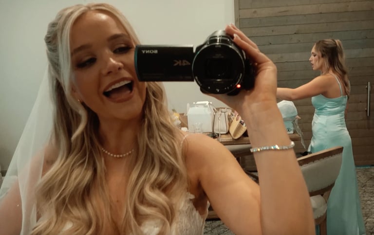 Woman in white dress holding a professional camera, smiling while another woman in teal poses in the background.