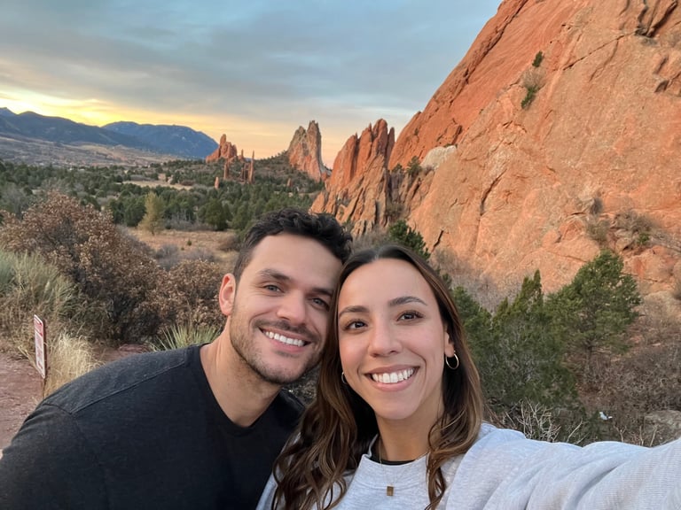 Smiling couple posing for selfie with red rock formations and mountains in background at sunset