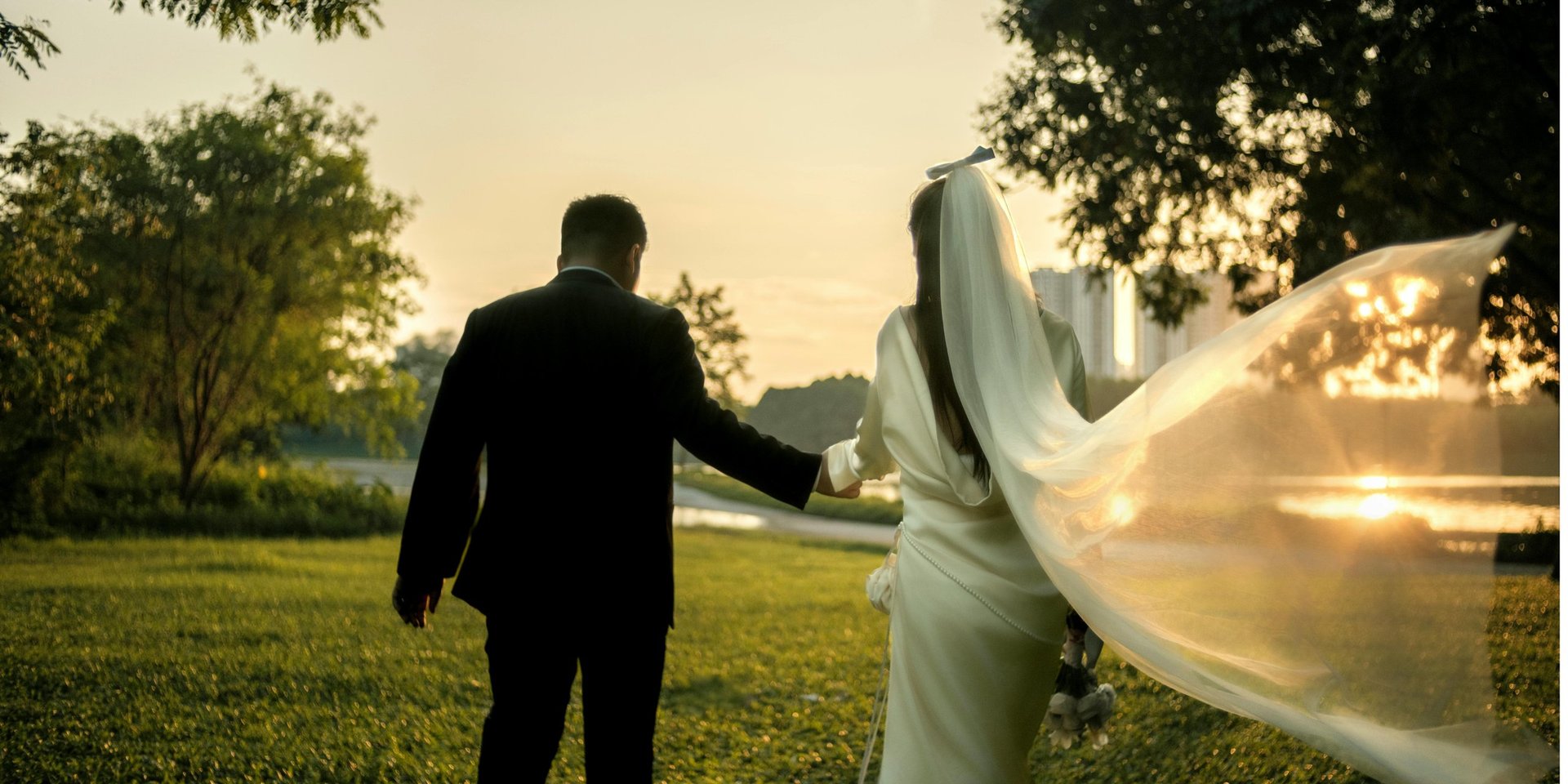 Bride and groom holding hands in a grassy field at sunset, with the brides veil flowing in the wind and trees in the background.