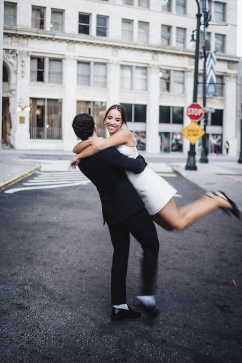 Woman in black and white outfit playfully lifting man on urban street with traffic signs and building backdrop